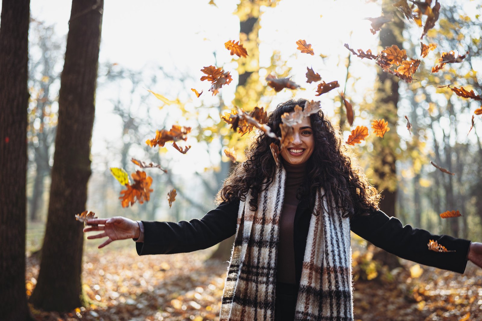 Backlit lifestyle photo of a woman with arms raised throwing maple leaves, celebrating autumn in a park.